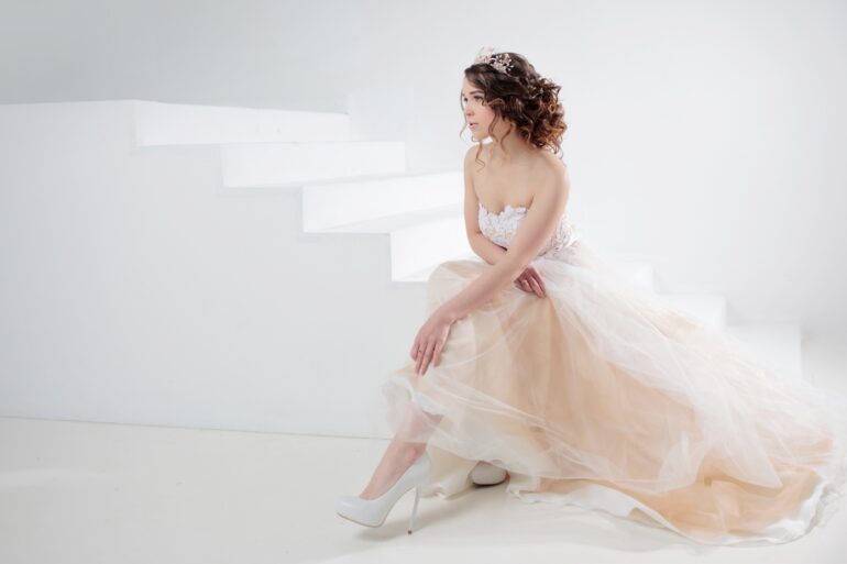 A woman wearing a pale wedding dress sitting on white stairs, with one leg extended and curled hair pinned back.
