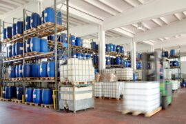 A warehouse is full of pallet rack system, and on those pallet racks are blue containers. There is a forklift in the foreground.