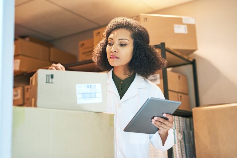 A female pharmacist is holding a tablet in one hand while she inspects a small cardboard box with the other.