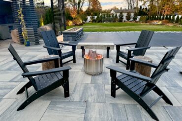 A large concrete patio in a residential backyard. Four chairs, two stools, and a bench on the patio surround a fire pit.