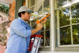 A man in a hat is using a chalk gun to apply chalk to an exterior window. He is standing on a ladder to apply the chalk.