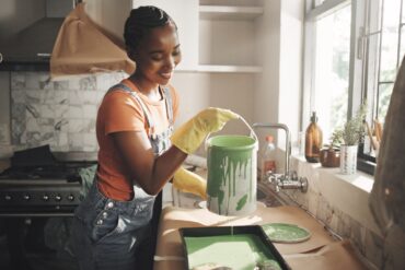 A woman in her home working on painting her kitchen on her own. She is pouring a bucket of green paint into a bin.