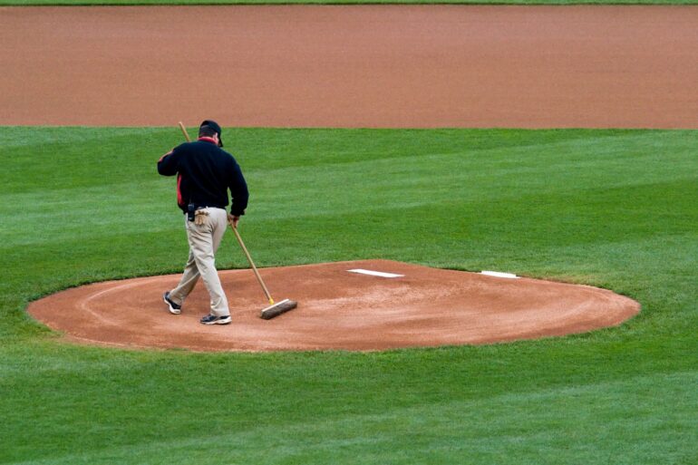 A person wearing tan pants, a black jacket, and a black baseball cap is using a brush to smooth a pitching mound.