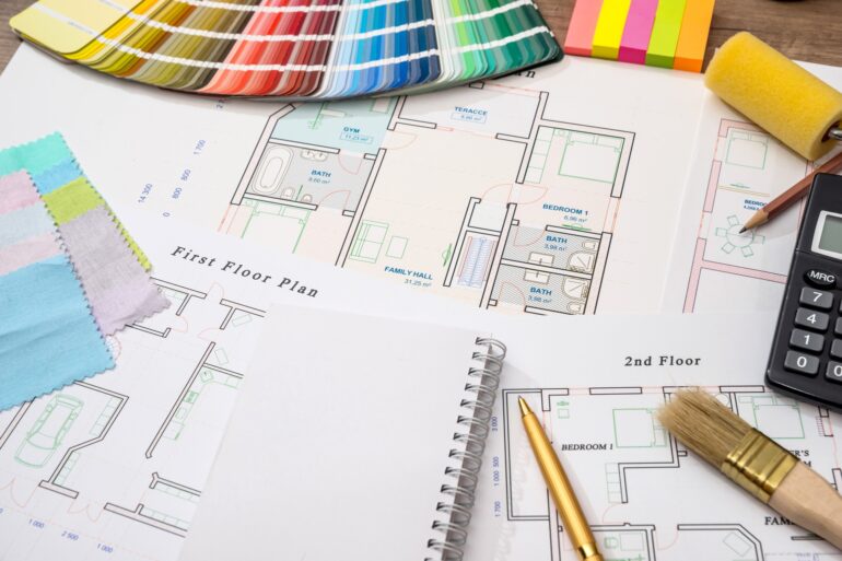 A calculator, pen, color swatches, and paintbrush resting on a house plan with printed room outlines.