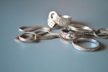 A braided silver chain and several silver rings with textured details resting on a soft blue surface.