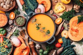 A top view of a pot of orange soup topped with seeds and herbs, surrounded by vegetables and kitchen utensils on wood.