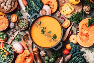 A top view of a pot of orange soup topped with seeds and herbs, surrounded by vegetables and kitchen utensils on wood.