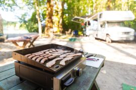 A camp griddle cooking strips of bacon and sausages on a picnic table with a recreational vehicle in the background.