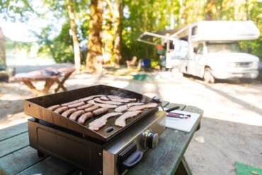 A camp griddle cooking strips of bacon and sausages on a picnic table with a recreational vehicle in the background.