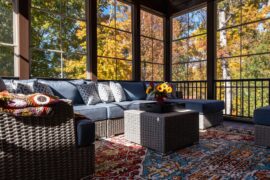 a sunroom with blue-cushioned wicker seating, a square table with flowers, tall windows, and autumn trees outside.