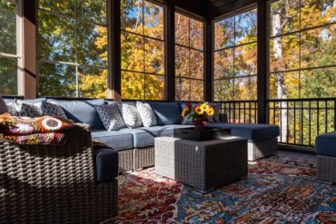 a sunroom with blue-cushioned wicker seating, a square table with flowers, tall windows, and autumn trees outside.