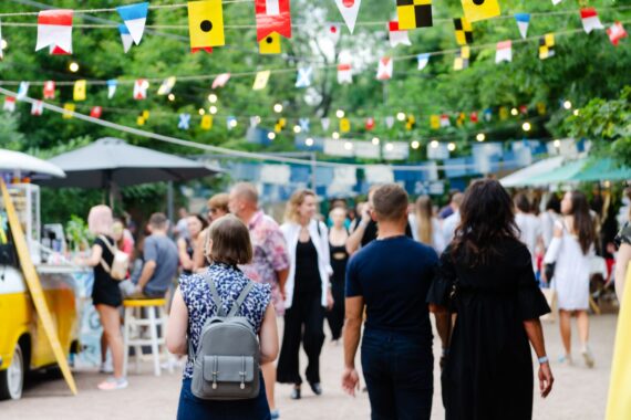 A crowd walking through an outdoor food festival with vendor tents, hanging flags, string lights, and trees.