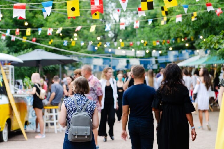 A crowd walking through an outdoor food festival with vendor tents, hanging flags, string lights, and trees.