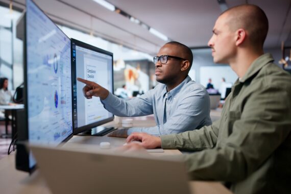 Two business men looking at two different computer monitors full of data. One is pointing at the screen.