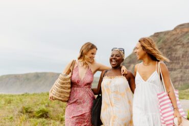 Three friends in summer dresses laugh together while walking through a coastal landscape with large tote bags.