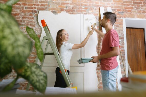A man and a woman are standing next to a brick wall. There is a wood piece against the wall they are painting white.