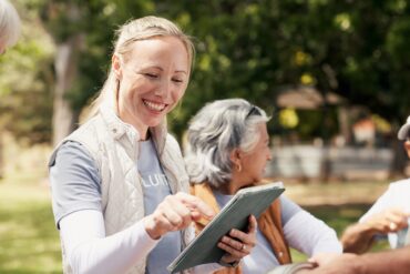 A volunteer smiles while using a tablet with neighbors during a community event in a sunny neighborhood park.