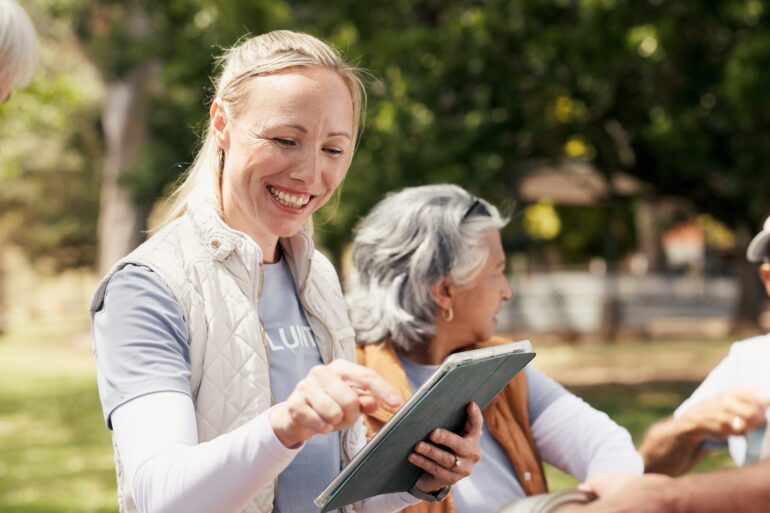 A volunteer smiles while using a tablet with neighbors during a community event in a sunny neighborhood park.