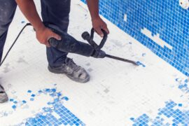 Worker uses a jackhammer to remove damaged blue mosaic tiles from the floor of an empty swimming pool during renovation.
