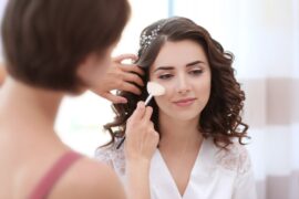 Bride with curled brown hair sits in a white robe as a makeup artist applies blush to her cheek with a brush.