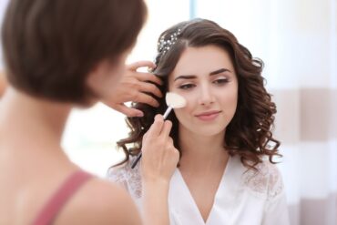 Bride with curled brown hair sits in a white robe as a makeup artist applies blush to her cheek with a brush.