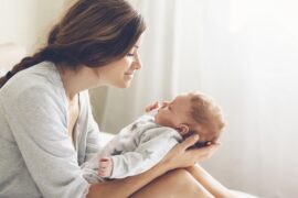 Mother smiles down at her newborn baby while gently cradling the infant in her arms beside a bright window.