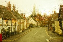 A rain-spattered window blurs a narrow street lined with buildings, parked cars, and warm lights under a pale sky.