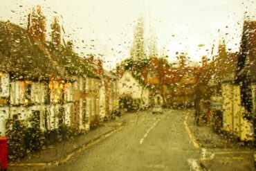 A rain-spattered window blurs a narrow street lined with buildings, parked cars, and warm lights under a pale sky.