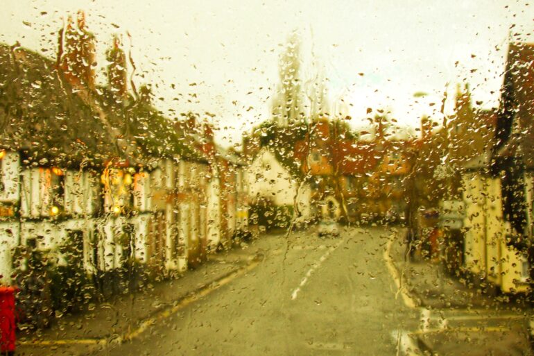 A rain-spattered window blurs a narrow street lined with buildings, parked cars, and warm lights under a pale sky.