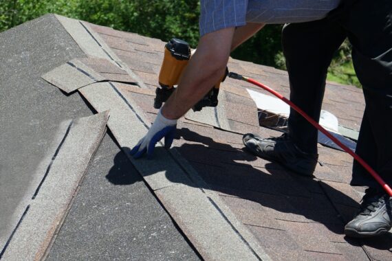 Roofer installs asphalt shingles with a pneumatic nail gun while working on a sloped residential roof.