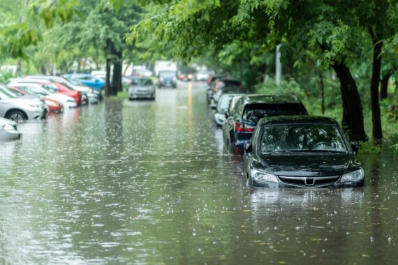 A residential street is lined with different parked vehicles. Floodwaters fill the street, and trees line the road.