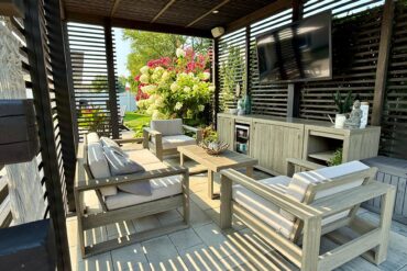 An outdoor space with a large wooden pergola over multiple couches, a coffee table, and a TV surrounded by greenery.