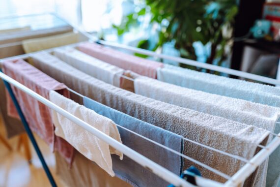Folding drying rack holds rows of damp towels and cloths in soft neutral and pastel colors inside a bright room.