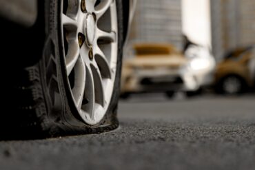 A dark gray car with a flat tire parked on the pavement outside. Two yellow cars are parked in the distance.