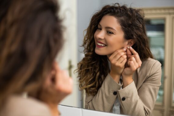 A smiling woman wearing a beige blazer is putting on an earring while looking into a mirror indoors.