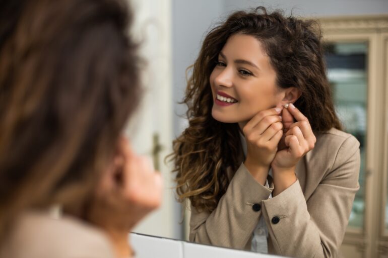A smiling woman wearing a beige blazer is putting on an earring while looking into a mirror indoors.