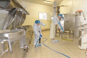 A man and a woman wearing protective suits and hairnets are cleaning the floor of an industrial space.