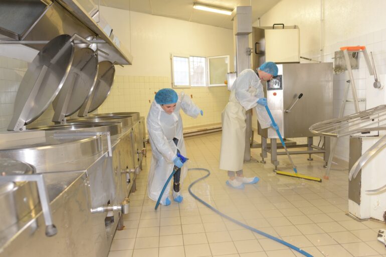 A man and a woman wearing protective suits and hairnets are cleaning the floor of an industrial space.